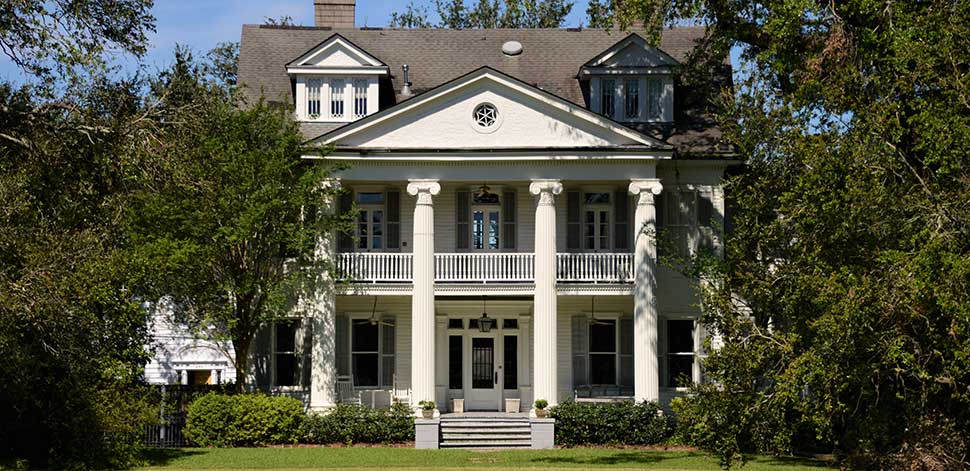 Large white two-story Louisiana home with elegant pillars and a balcony, nestled among native trees under a bright blue sky.
