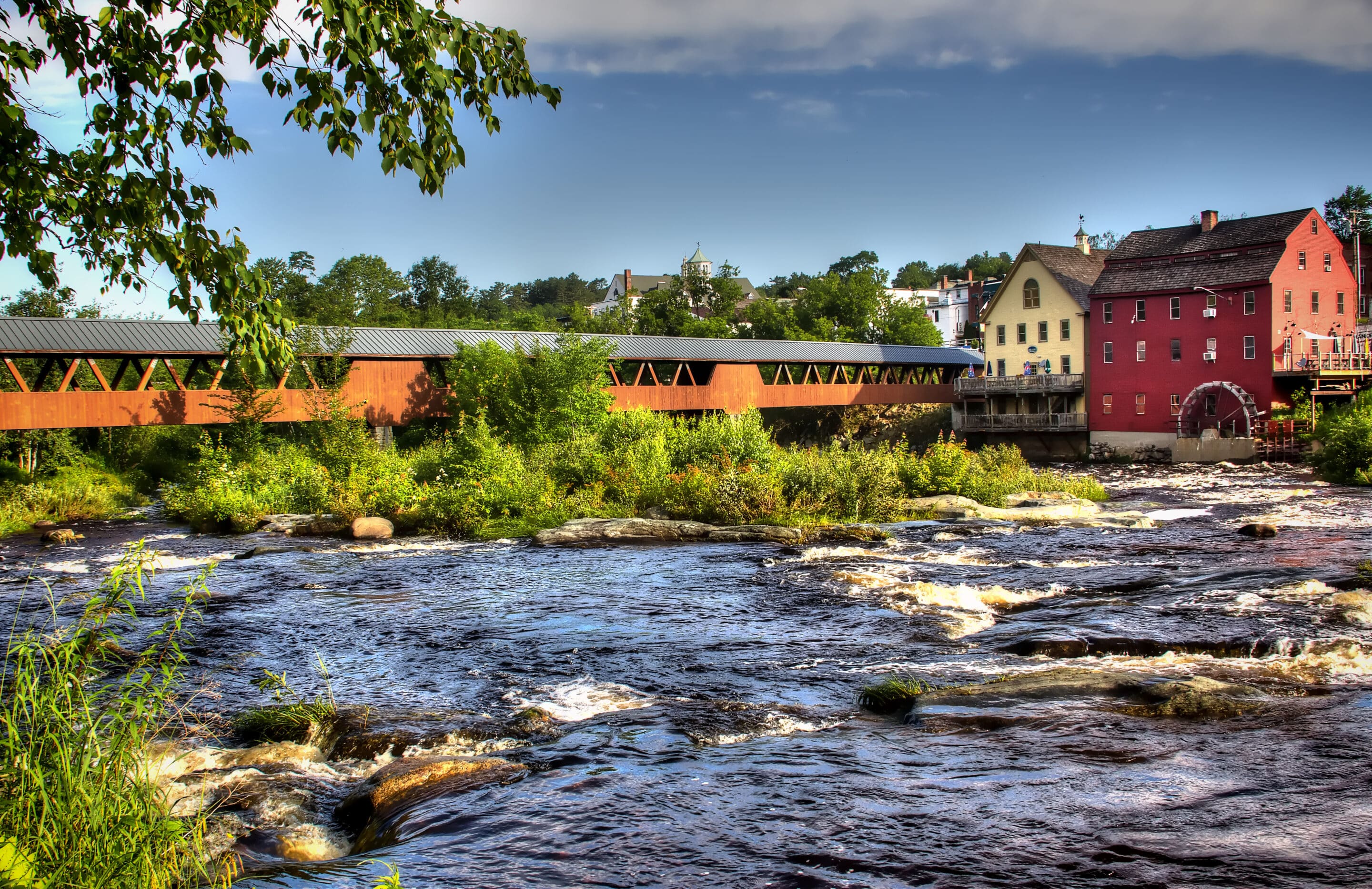 Bridge over the Ammonoosuc River with a grist mill and foliage, blue skies, and a view of Littleton, NH in the distance.