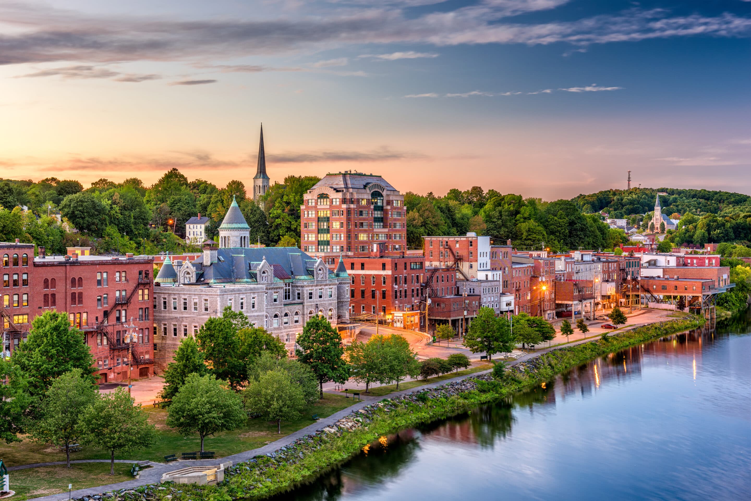 A Maine riverside town at dusk, reflecting the importance of Maine flood insurance against the backdrop of historic buildings and a gentle river, with lights beginning to twinkle in the evening tranquility.
