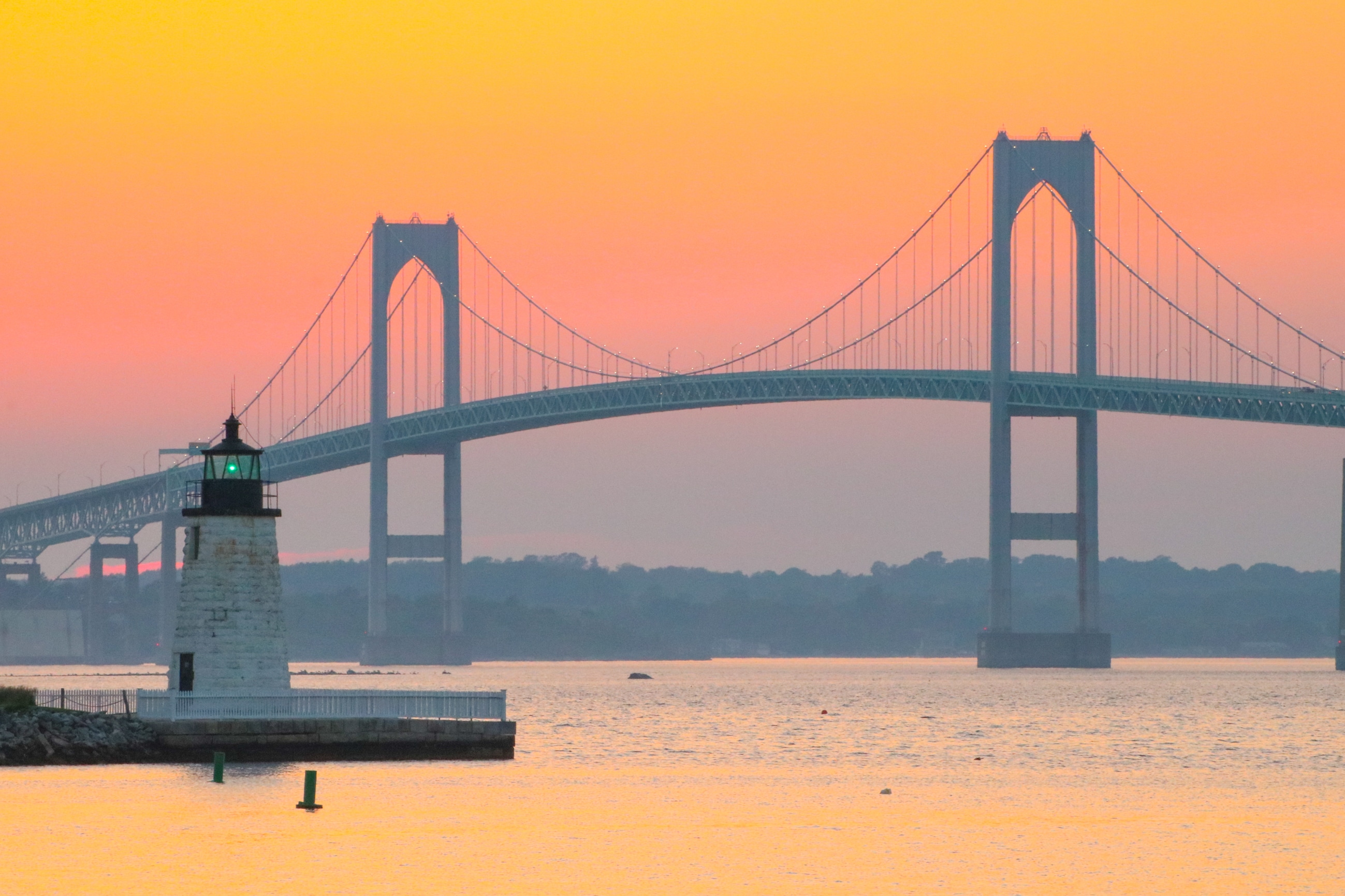 A Rhode Island lighthouse and suspension bridge bathed in the orange-pink light of sunset, symbolizing the protection offered by Rhode Island flood insurance in coastal communities.