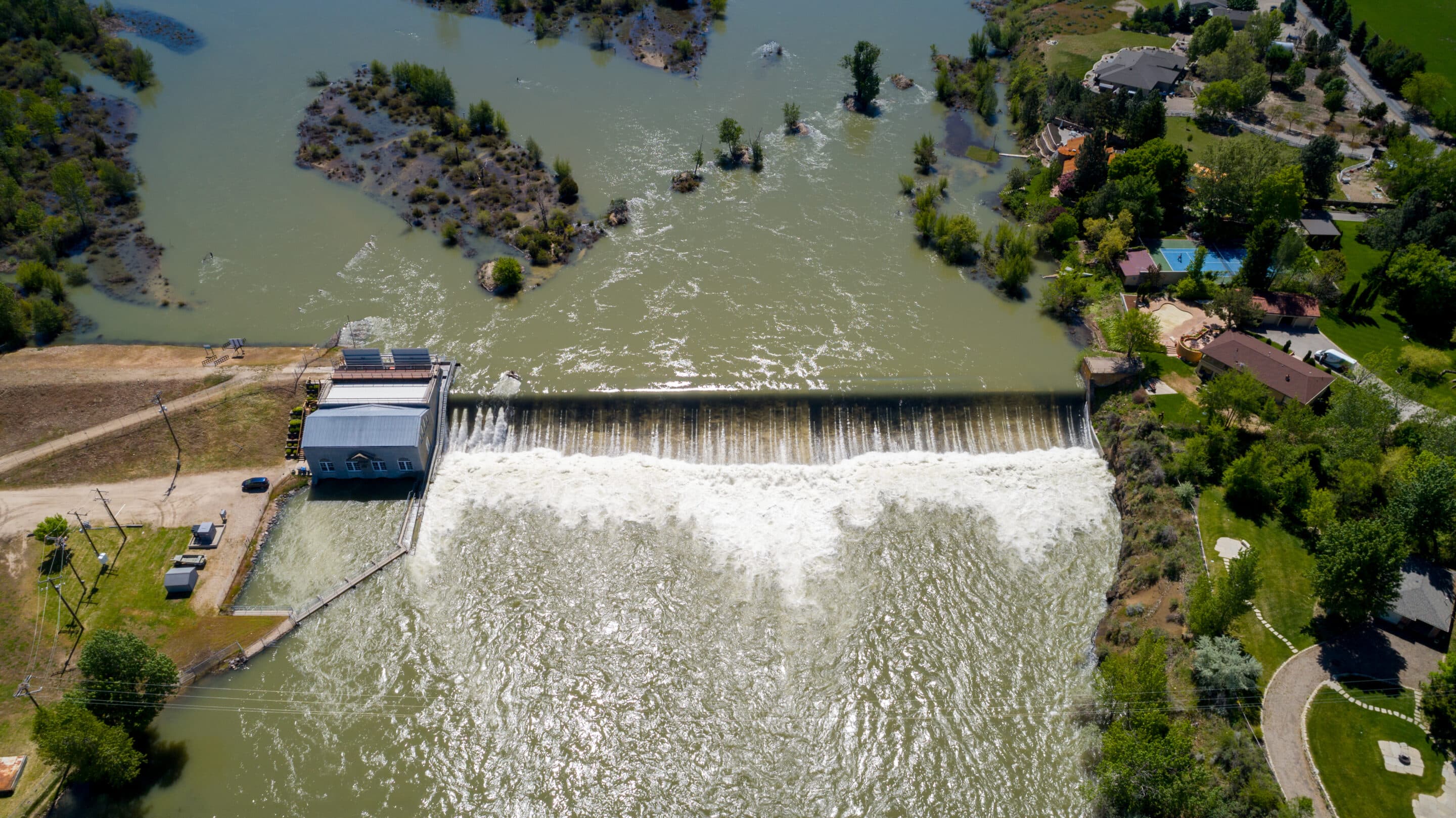 Aerial view of a dam releasing water near suburban homes and green landscape in Idaho.