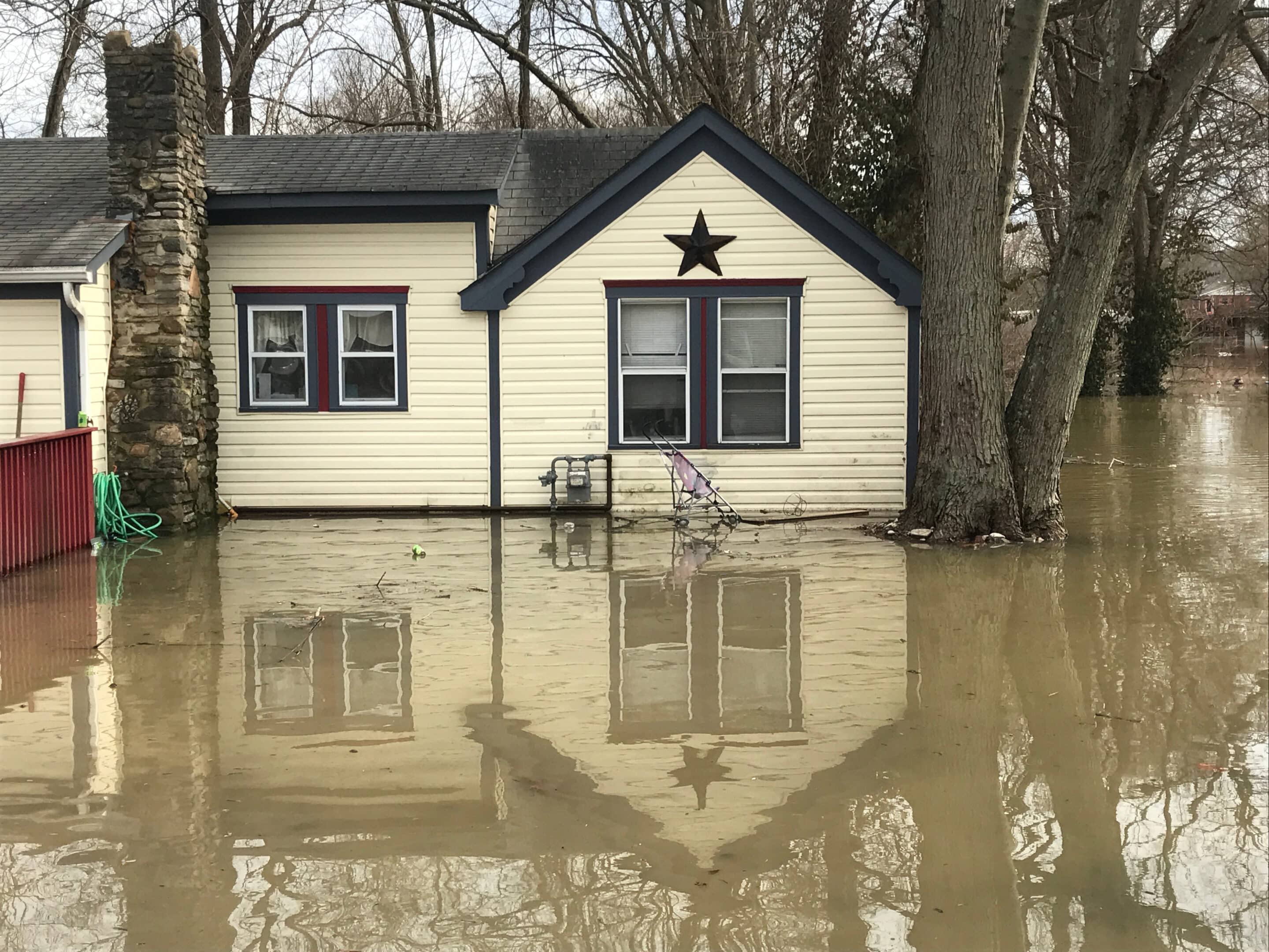 Floodwaters rise around a single-story house with trees and brown water reflecting the home exterior.