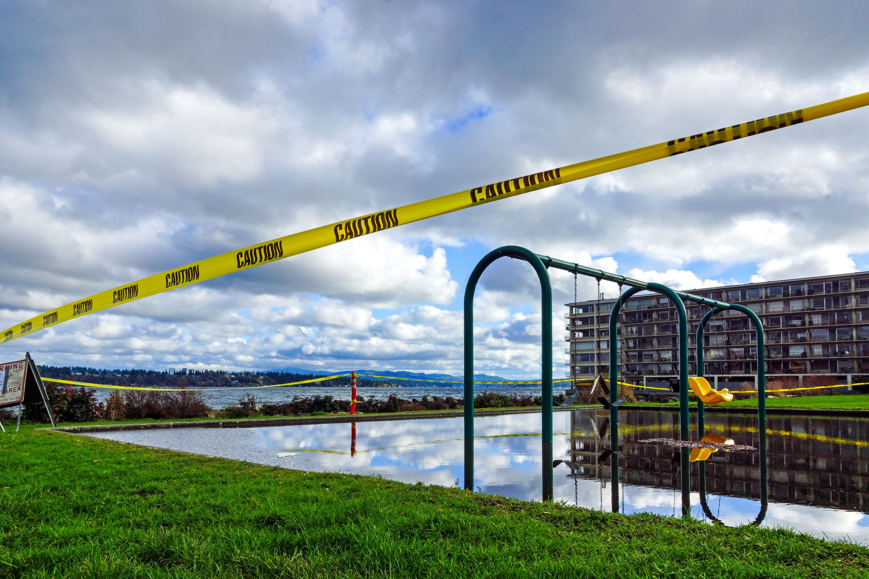 Flooded playground surrounded by caution tape near residential buildings and waterfront.