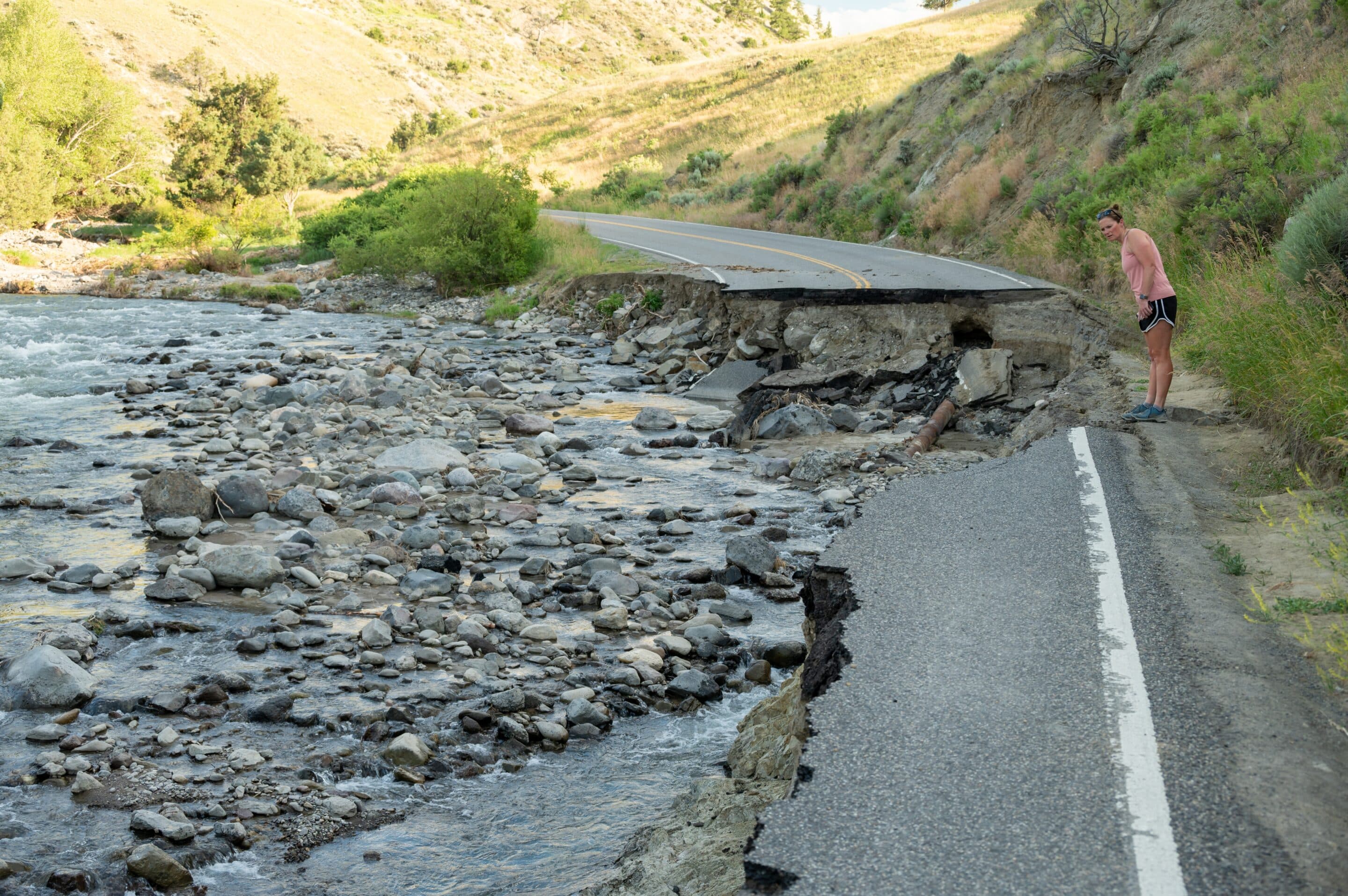 Collapsed rural road next to a flooded rocky riverbed with a person standing on the edge.